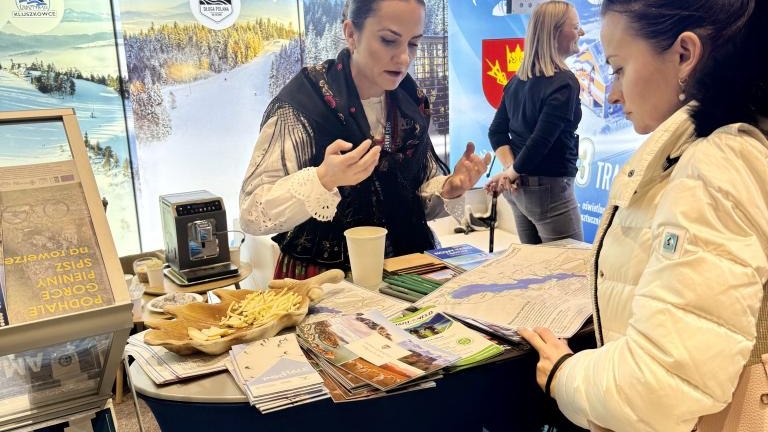 Dwie kobiety rozmawiąjące ze sobą na wydarzeniu promocyjnym na tle podświetlonego baneru, między nimi stolik z ulotkami..jpeg