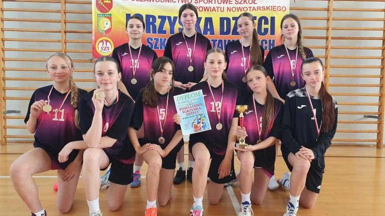 Oficjalne zdjęcie drużyny w fioletowo-czarnych strojach, zapozowane na tle bramki do piłki ręcznej. Dziewczęta klęczą i stoją, prezentując puchar, dyplom oraz medale. Przed nimi na parkiecie leżą dwie piłki sportowe oraz puchar.