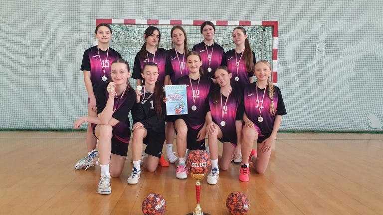 Oficjalne zdjęcie drużyny w fioletowo-czarnych strojach, zapozowane na tle bramki do piłki ręcznej. Dziewczęta klęczą i stoją, prezentując puchar, dyplom oraz medale. Przed nimi na parkiecie leżą dwie piłki sportowe oraz puchar.