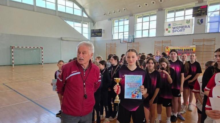 Zbliżenie na halę sportową. Starszy mężczyzna w bordowej bluzie sportowej stoi obok młodej zawodniczki w fioletowej koszulce z numerem 12. Dziewczyna z uśmiechem trzyma w rękach puchar i dyplom „POWIATOWE IGRZYSKA DZIECI”. W tle widać pozostałe uczestnicz