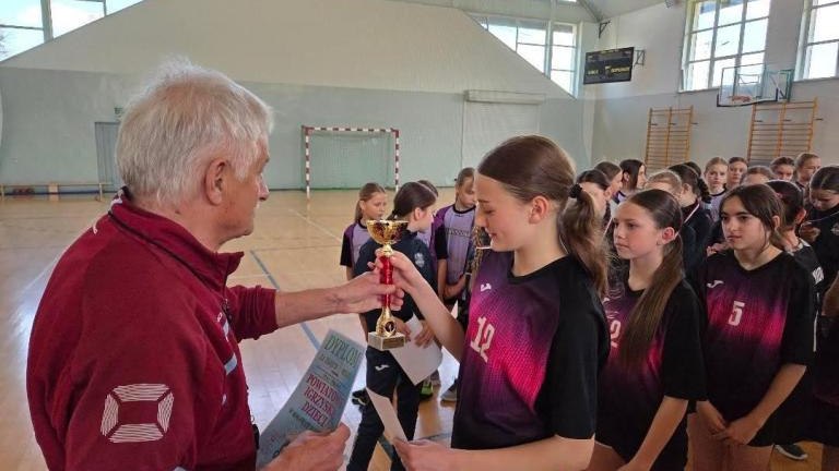 Ujęcie z bliska przedstawiające moment, w którym starszy mężczyzna (trener lub organizator) uroczyście przekazuje puchar kapitance drużyny. Dziewczyna jest ubrana w fioletowy strój sportowy, a w tle widać skupione twarze jej koleżanek z zespołu czekającyc