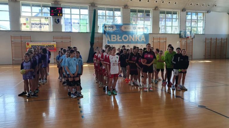 Szerokie ujęcie hali sportowej wypełnionej grupami dzieci w różnokolorowych strojach sportowych (fioletowych, błękitnych, czerwonych, czarnych). Zespoły stoją w rzędach na parkiecie podczas oficjalnego otwarcia. W tle widoczne duże okna, drabinki gimnasty