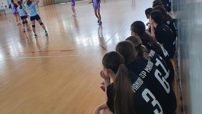 Widok z boku hali sportowej. Na pierwszym planie, na drewnianej ławie, siedzą tyłem zawodniczki w czarnych koszulkach z napisem „MANIOWY”. W głębi, na środku boiska, trwa mecz lub rozgrzewka – widać dziewczęta w błękitnych i fioletowych strojach. Na ścian