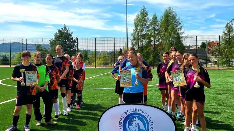 Trzy grupy młodych piłkarek w różnych strojach sportowych (czarnych, błękitnych i fioletowych) pozują z pucharami i dyplomami na środku boiska. Przed nimi widoczne logo.jpeg