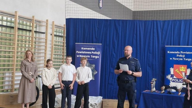 Scena z ceremonii wręczenia nagród. Policjant w okularach przemawia do mikrofonu, trzymając kartkę. Obok niego stoi policjantka z pucharami, a po lewej stronie widać troje dzieci z opiekunką.