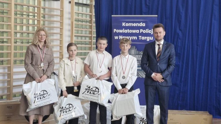 Troje dzieci z medalami i pucharami stoi w rzędzie wraz z dwojgiem dorosłych opiekunów. Wszyscy trzymają białe torby z nagrodami. W tle widoczne logo Komendy Powiatowej Policji w Nowym Targu.