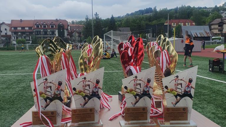 Puchary i statuetki lekkoatletyczne ustawione na stole na tle boiska.