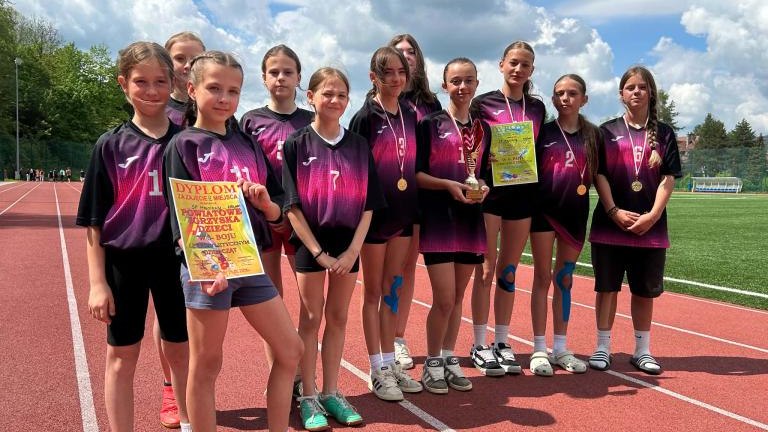 Drużyna w czarno-fioletowych strojach pozuje na bieżni z medalem, pucharem i dyplomem za pierwsze miejsce.