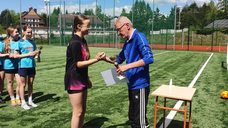 Moment dekoracji na boisku sportowym. Mężczyzna w niebieskiej bluzie sportowej i czarnych spodniach podaje rękę zawodniczce w czarno-różowym stroju, trzymając w drugiej dłoni puchar i dyplom.