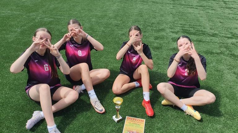 Cztery zawodniczki w czarno-różowych strojach siedzą na murawie boiska, wykonując palcami zabawne gesty wokół oczu. Przed nimi na trawie leży dyplom i stoi sportowe trofeum.