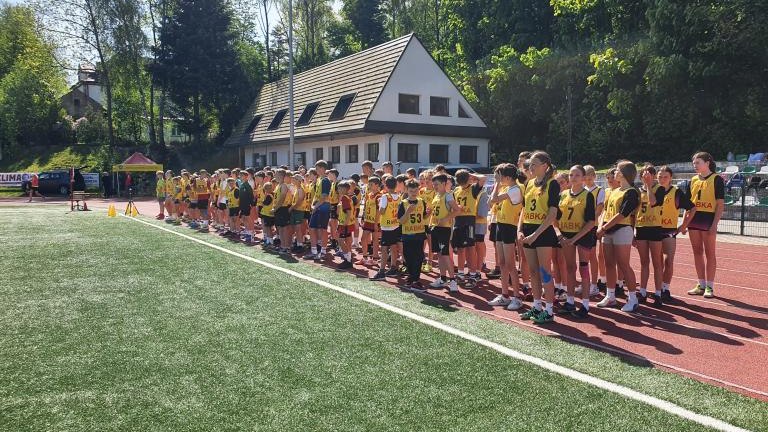 Szerokie ujęcie przedstawiające dużą grupę sportowców stojących w długim rzędzie na czerwonej bieżni lekkoatletycznej, wzdłuż zielonej murawy boiska. Wielu uczestników ma na sobie żółte koszulki z napisem „Rabka”. W tle widoczny jest biały budynek z ciemn