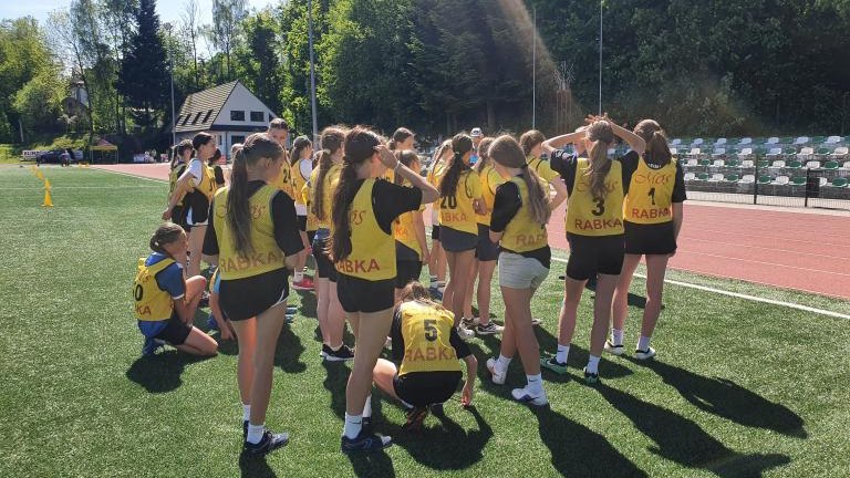 Grupa zawodników w żółtych znacznikach sportowych z napisem „Rabka” stoi w skupisku na zielonej murawie boiska, tuż obok czerwonej bieżni. Zdjęcie wykonane w pełnym słońcu, z widocznymi długimi cieniami na trawie.