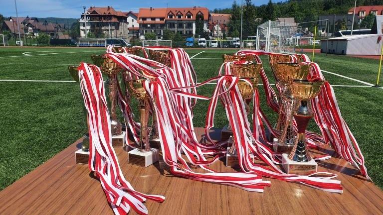 Zbliżenie na drewniany stół ustawiony na zielonej murawie boiska sportowego, na którym wyeksponowano rząd złotych pucharów. Każde trofeum jest udekorowane podłużnymi, biało-czerwonymi wstęgami. W tle widoczne są budynki mieszkalne i zielone wzgórza.