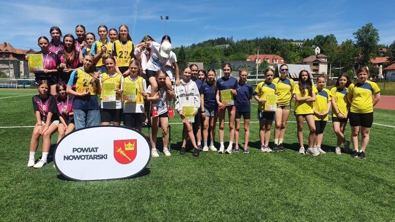 Pamiątkowe zdjęcie grupowe zawodniczek stojących na zielonej murawie boiska. Uczestniczki w różnych strojach sportowych prezentują zdobyte dyplomy i medale. Na pierwszym planie, na trawie, ustawiona jest biała owalna tablica z napisem „POWIAT NOWOTARSKI”