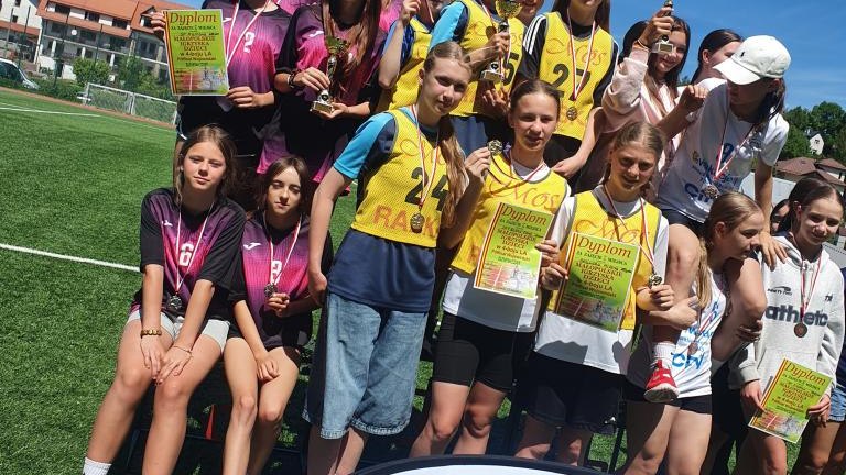 Bliższe ujęcie grupy zawodniczek pozujących wspólnie do zdjęcia na tle boiska. Dziewczęta trzymają w rękach trofea, medale oraz zielone dyplomy sportowe. Przed grupą widoczna jest dolna część tablicy z napisem „POWIAT NOWOTARSKI”.