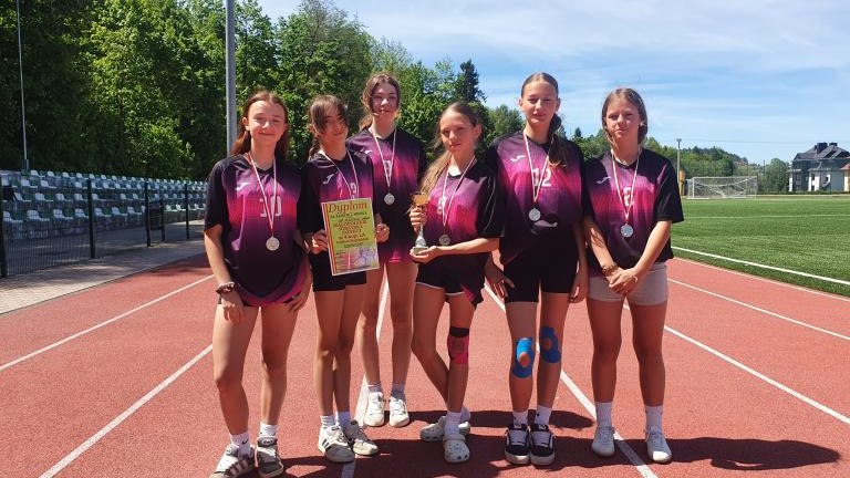 Sześć zawodniczek w jednakowych, ciemnoróżowo-czarnych koszulkach sportowych stoi obok siebie na czerwonej bieżni lekkoatletycznej. Dziewczęta mają na szyjach srebrne medale, a dwie z nich trzymają w dłoniach puchar oraz pamiątkowy dyplom zawodów.