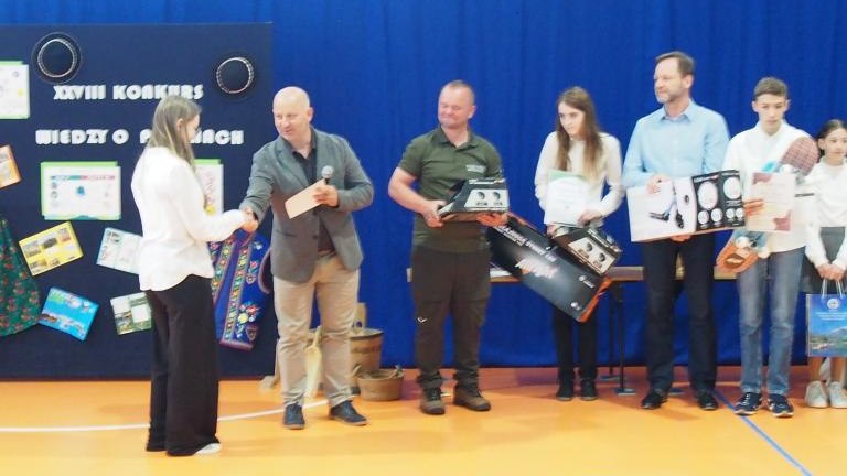 Moment wręczania nagród podczas „XXVIII Konkursu Wiedzy o Pieninach”. Mężczyzna w szarej marynarce podaje rękę stojącej przed nim uczennicy. W tle pozostali dorośli oraz uczniowie trzymają nagrody rzeczowe, w tym zapakowane hulajnogi i dyplomy.