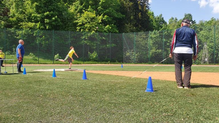 Szerokie ujęcie rzutni lekkoatletycznej z zawodnikiem kończącym pchnięcie kulą oraz sędzią z taśmą mierniczą.