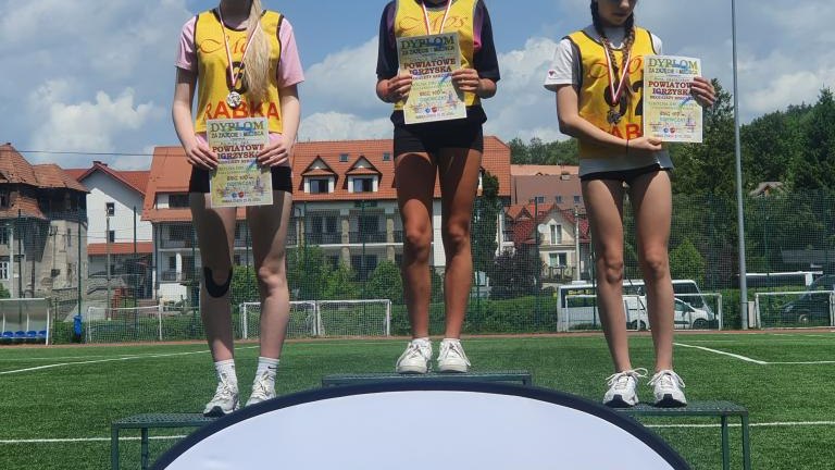 Trzy zawodniczki z dyplomami stoją na podium, przed którym znajduje się duży szyld z napisem „Powiat Nowotarski”.