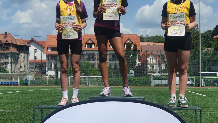 Trzy lekkoatletki z medalami i dyplomami pozują na podium za oficjalną tablicą Powiatu Nowotarskiego.