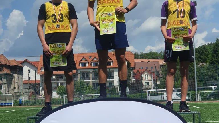 Trzej najlepsi zawodnicy zawodów lekkoatletycznych stoją na podium z medalami i dyplomami.