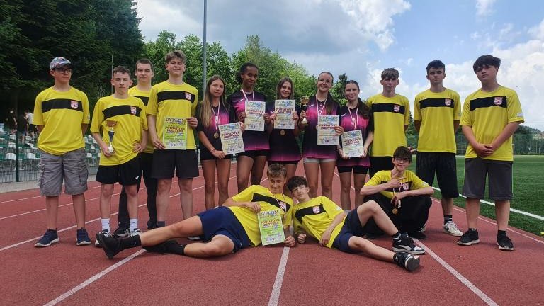 Wspólne, radosne zdjęcie grupowe sportowców z pucharami i dyplomami na czerwonej bieżni lekkoatletycznej.